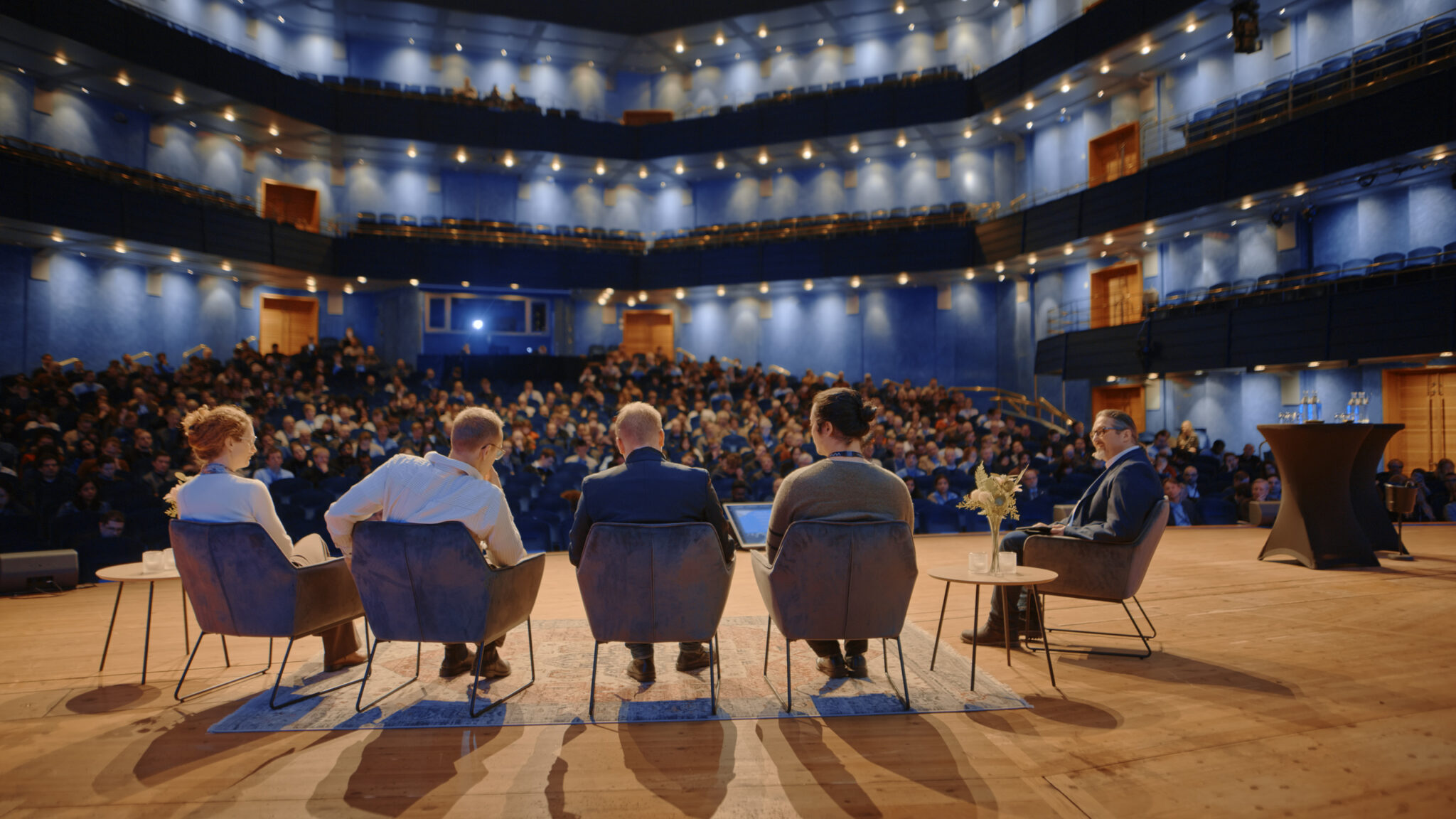 Blue theatre seen from the stage with five people in chairs in the foreground.